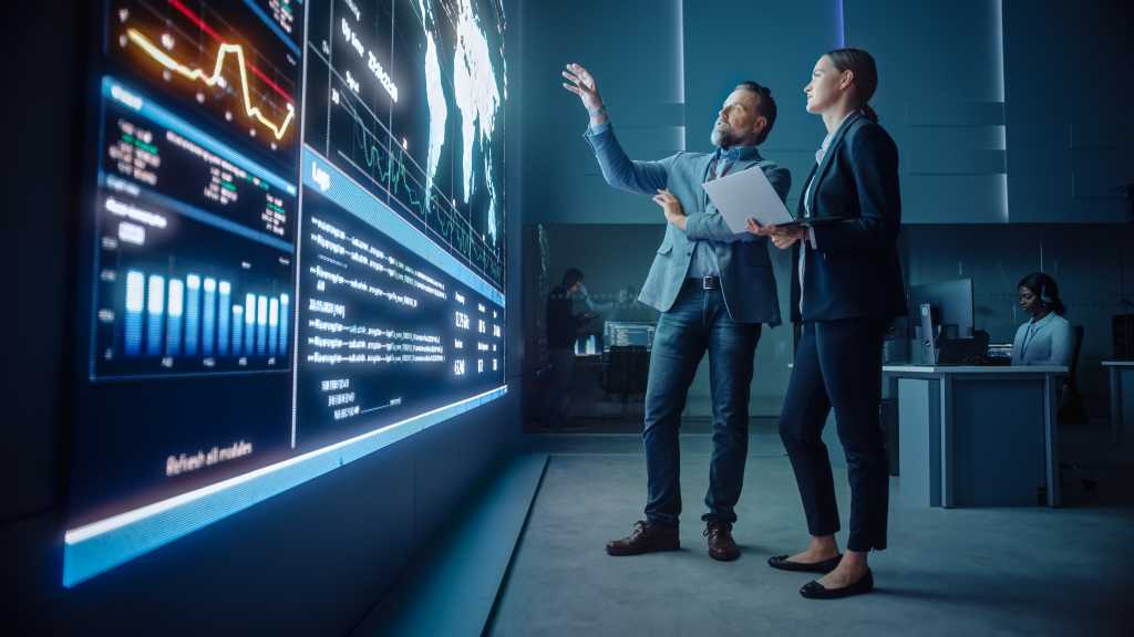 Project Manager and Computer Science Engineer Talk while Using Big Screen Display and a Laptop, Showing Infrastructure Infographics Data. Telecommunications Company System Control and Monitoring Room.