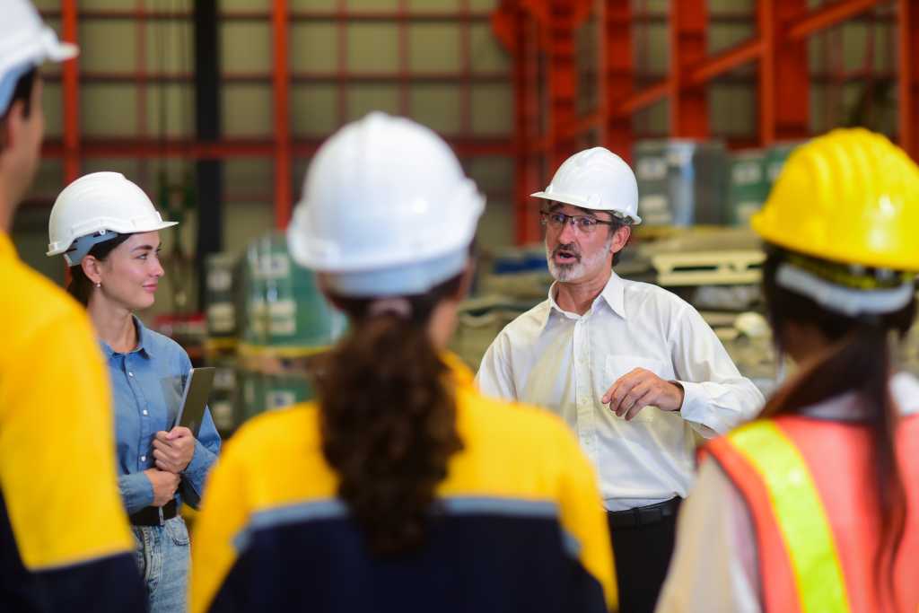 Professional Caucasian manager engineer wearing safety hardhat is training industry worker, workshop factory operator, mechanical engineering team production in heavy industrial manufacture factory.