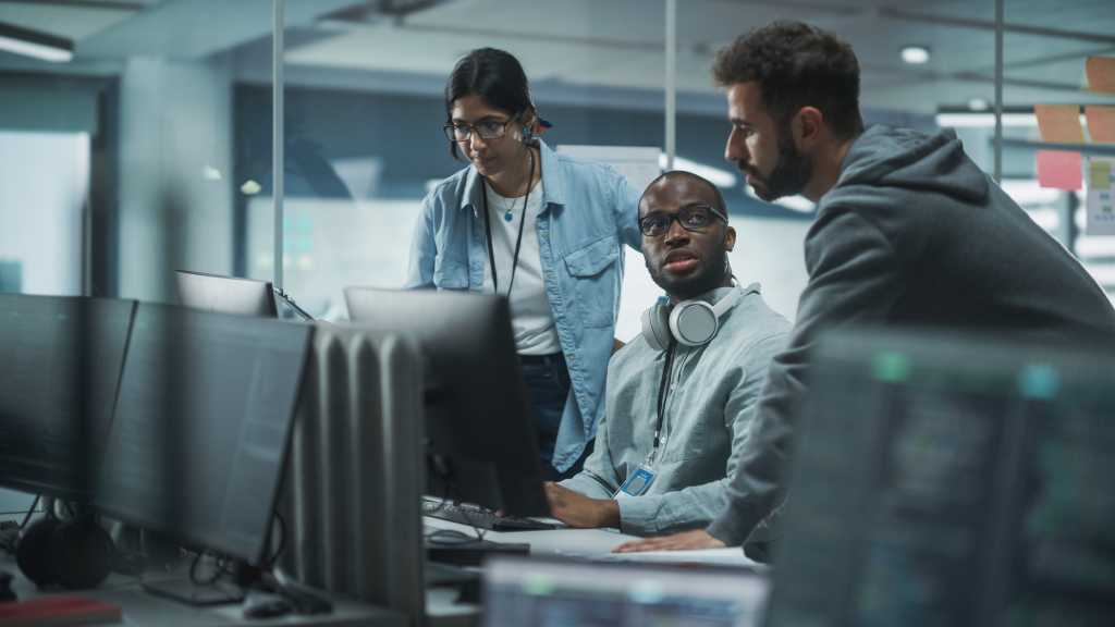 Diverse Multi-Ethnic Team in Modern Office: Teamwork of IT Programmers Gather Around Desktop Computer, Talking, Finding Solution. Specialists Create Software. Engineers Develop App, Program